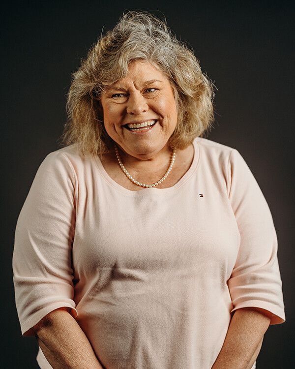 Woman with light brown hair, smiling, wearing a light pink sweater and a pearl necklace, against a dark background.