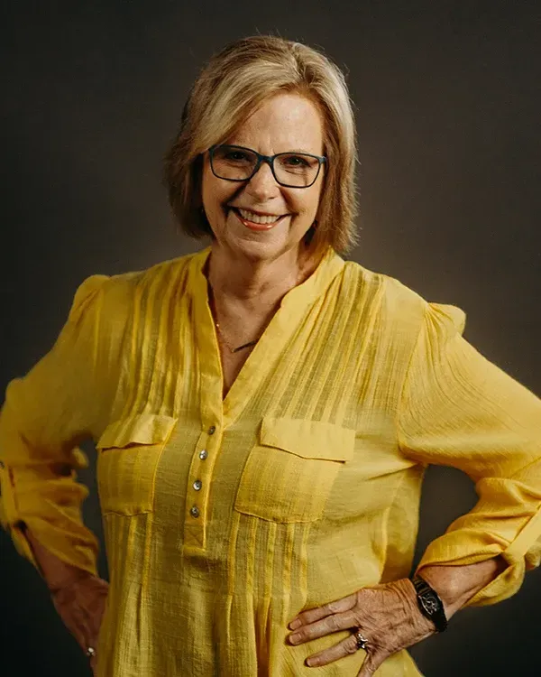 Woman in yellow shirt, glasses, and watch, smiling, hands on hips, against a gray background.