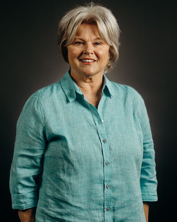 Woman with short, light hair wearing a turquoise button-down shirt smiles against a gray background.