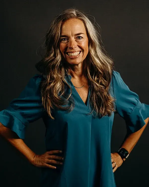 Woman with long gray hair smiles, wearing a teal blouse, hands on hips, against a dark background.