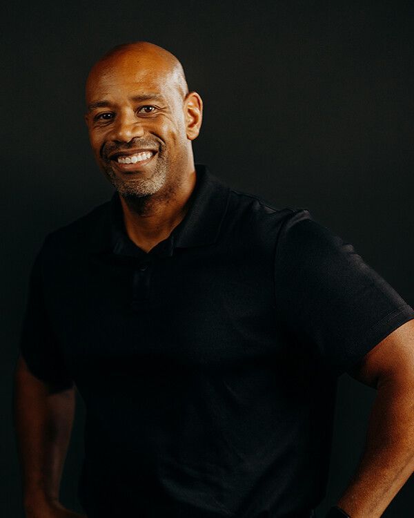 Man in black polo smiling against a dark background.