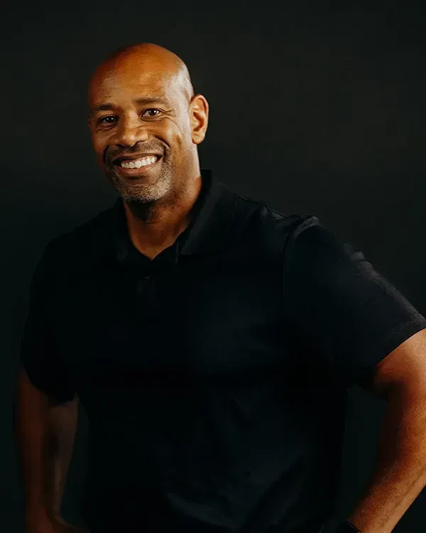 Man with a shaved head smiles, wearing a black polo shirt, against a dark background.