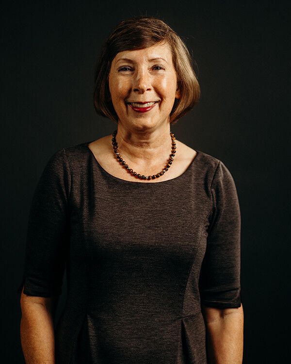 Woman with short brown hair smiles, wearing a dark dress and necklace against a dark background.