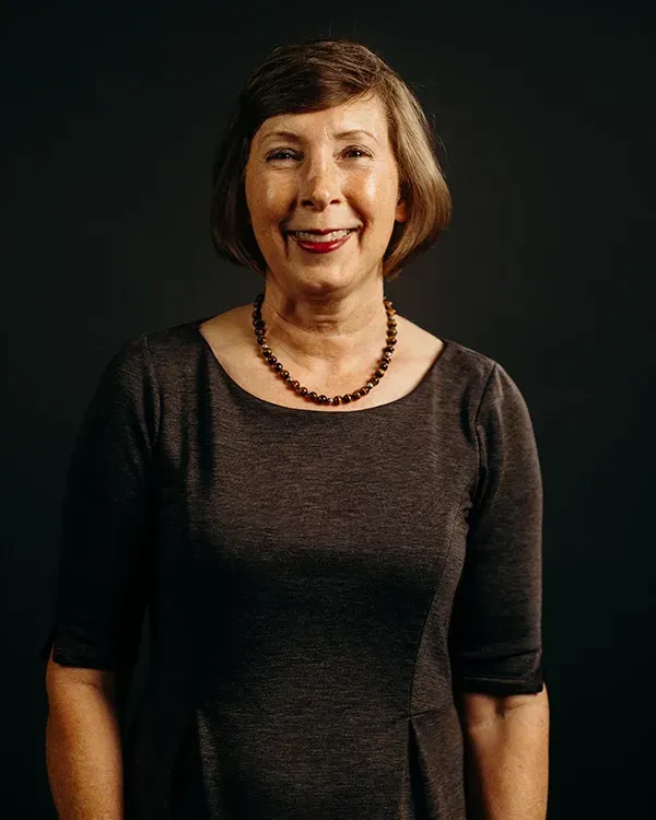 Woman with short hair smiles, wearing a dark brown dress and necklace against a dark background.
