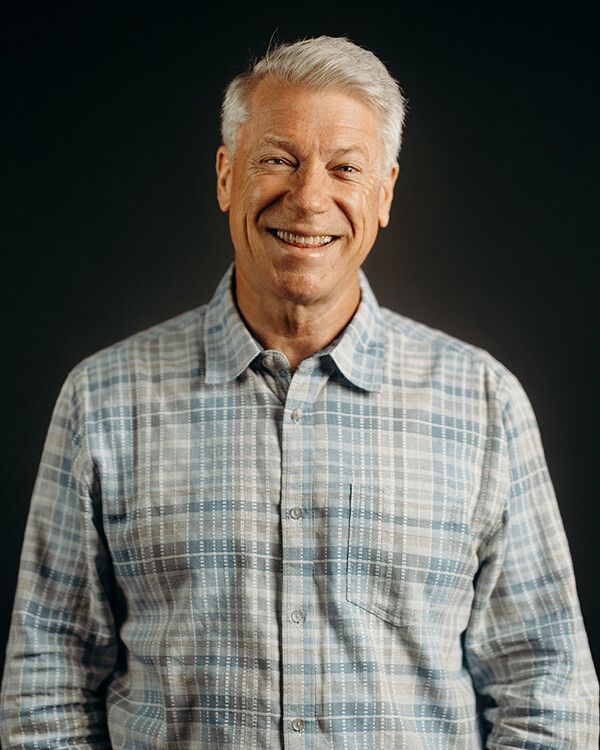 Man with grey hair and blue plaid shirt smiling, dark background.