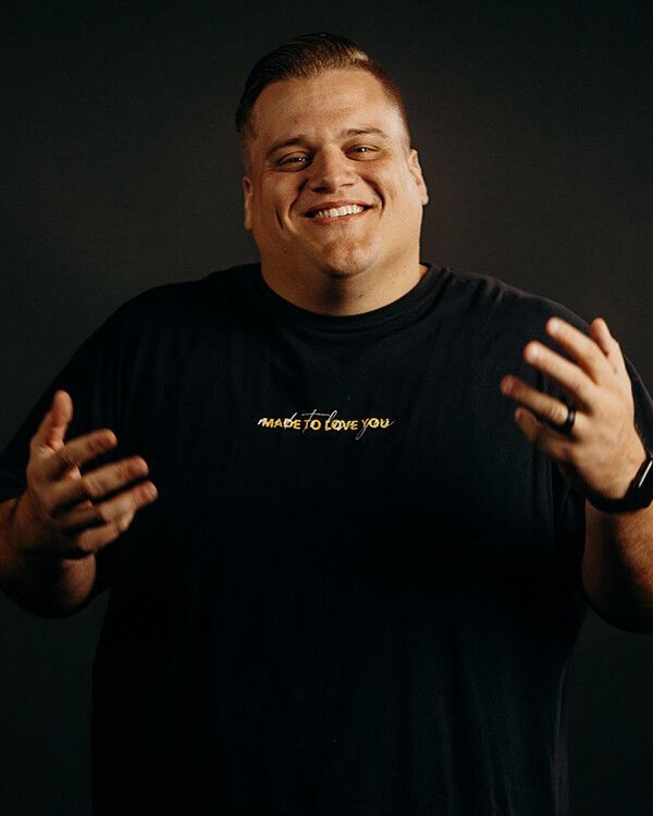 Man smiling, wearing a black t-shirt with gold text, hands open, against a dark backdrop.