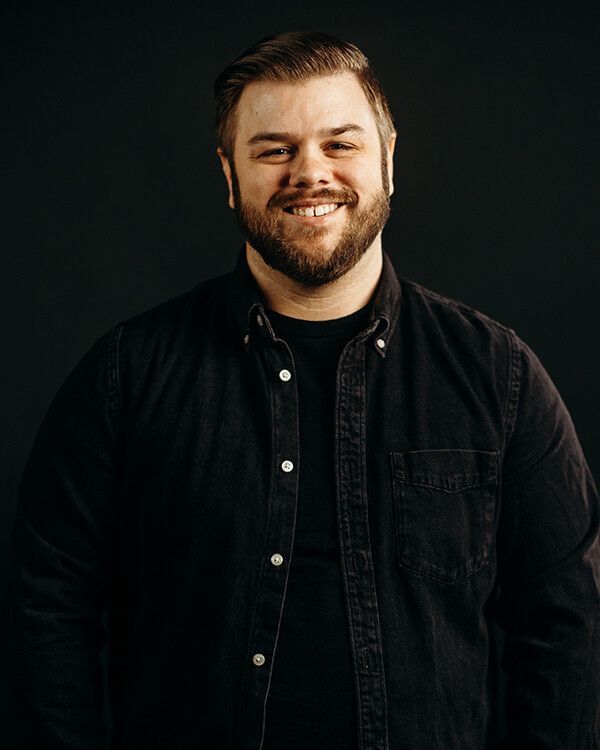Man with a beard smiling, wearing a dark button-down shirt, against a dark background.