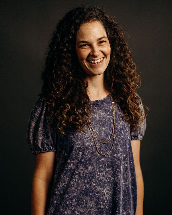 Woman with curly brown hair smiles, wearing a purple patterned top, long necklace against a dark background.