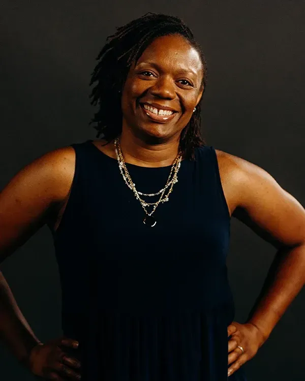 Woman in navy dress smiles, hands on hips. Dark background.