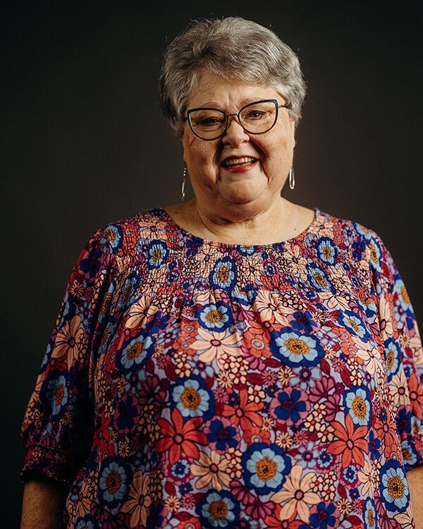 Woman in floral shirt and glasses smiles, against a dark backdrop.