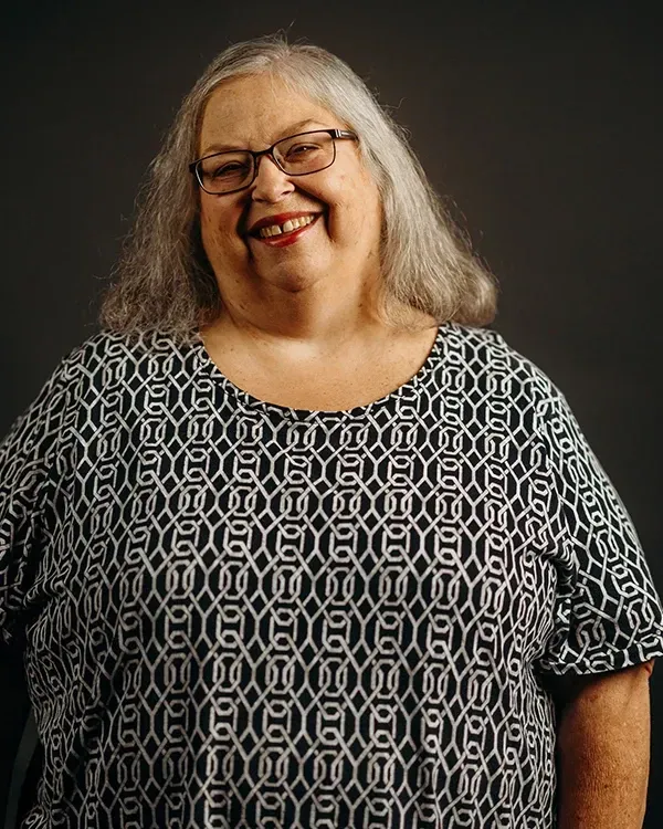 Woman with gray hair, glasses, and a patterned top smiles against a dark background.