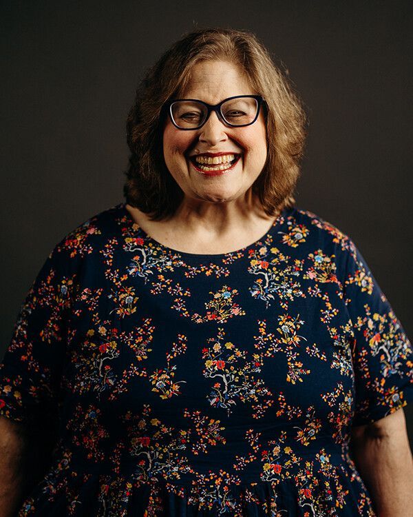 Woman wearing glasses smiles at the camera, dark blue dress with floral pattern, gray background.