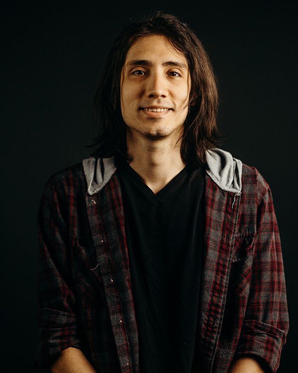 Person with long brown hair smiles, wearing black shirt, hooded top, and plaid shirt. Dark background.