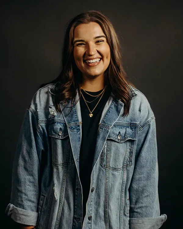 Woman in denim jacket smiling at the camera, wearing necklaces, against a dark gray background.