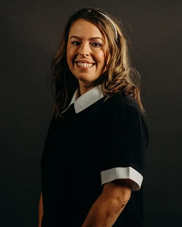 Woman smiles at the camera, wearing a black dress with white collar and cuffs, against a dark backdrop.