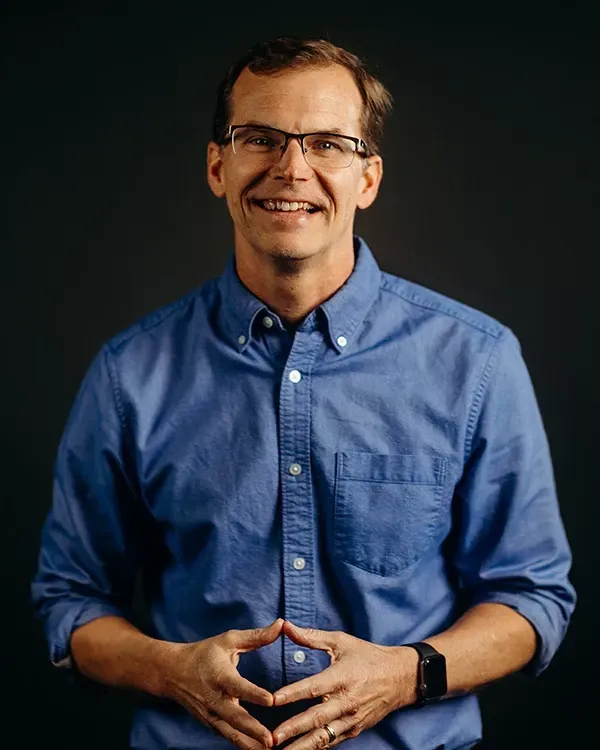 Man in glasses and blue shirt smiles, hands clasped, in front of a dark background.