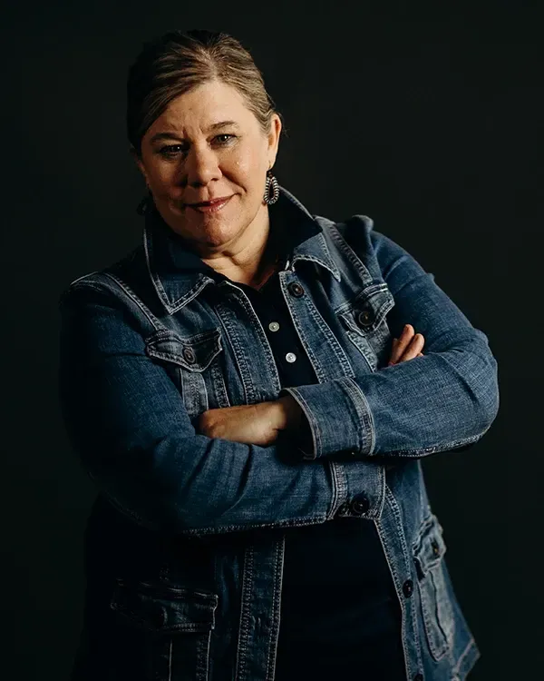 Woman with arms crossed, wearing a denim jacket and dark top, smiling slightly, against a dark backdrop.