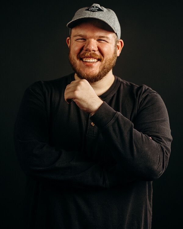 Man in gray cap and dark shirt smiles, hand to his chin, against a dark background.