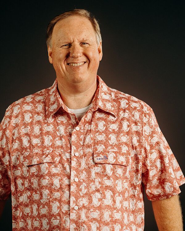Man smiling, wearing a red patterned button-down shirt, standing against a dark background.