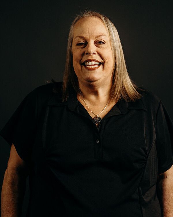 Woman with blonde hair smiling, wearing a black shirt, against a dark background.