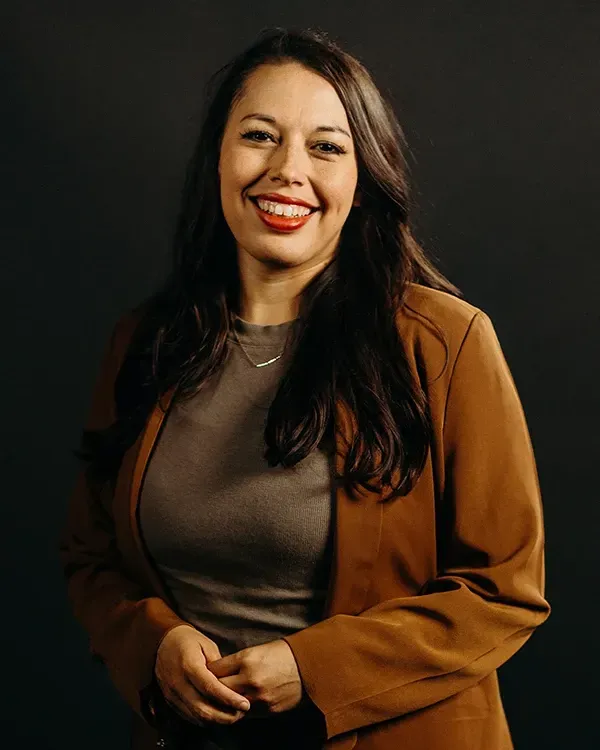 Woman with dark hair, wearing a brown jacket, smiling with red lipstick, looking at the camera.