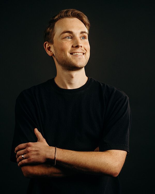 Man in black shirt, arms crossed, smiling against a dark backdrop.