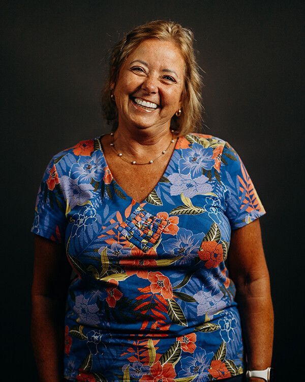Woman smiling, wearing a blue floral shirt, dark background.