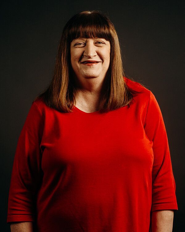 Woman in red shirt smiles at the camera, brown hair with bangs, against a dark background.
