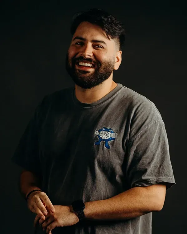 Man with dark hair and beard smiles, wearing a gray t-shirt with a small graphic, posing against a dark background.