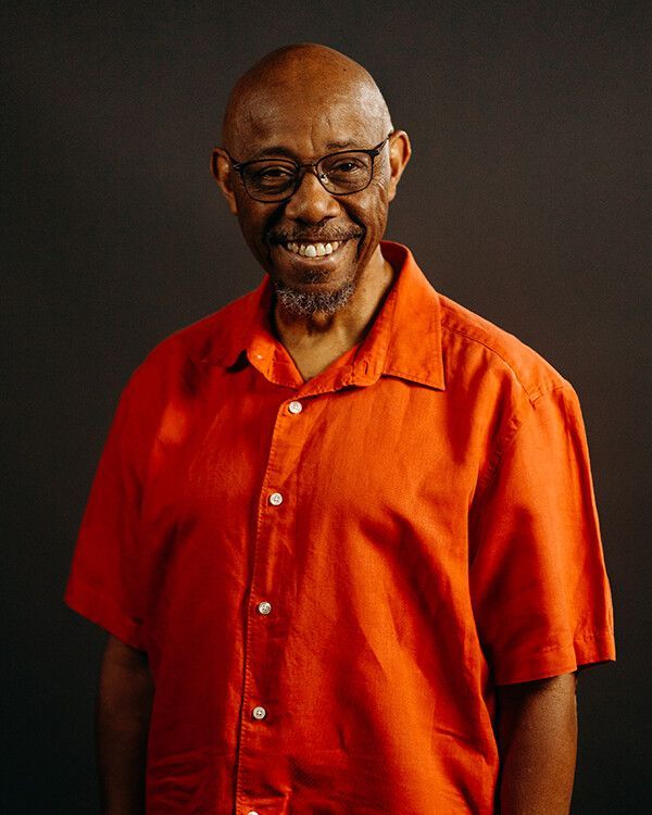 Man with glasses wearing a bright orange button-down shirt smiles, standing against a dark background.