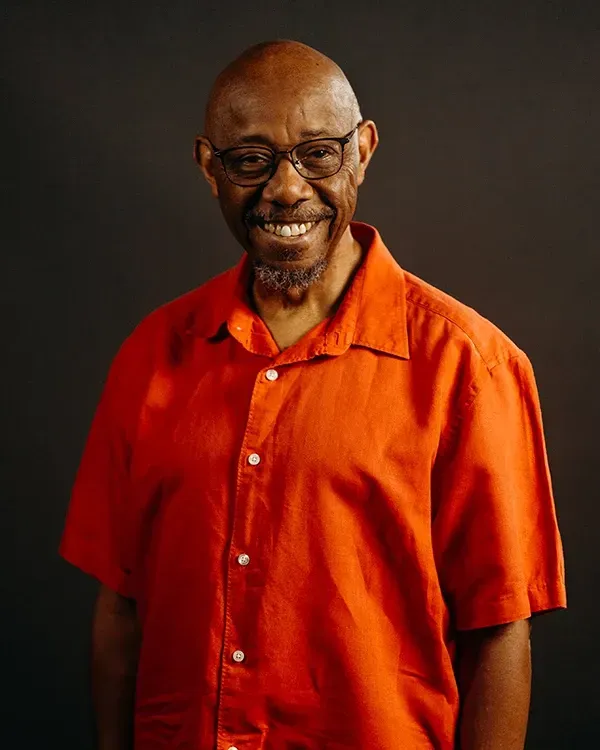Man with glasses smiling, wearing an orange button-down shirt, against a dark background.