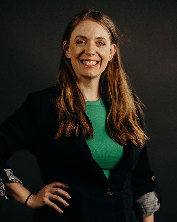 Woman with long brown hair, wearing a green top and black blazer, smiling with hand on hip. Dark background.