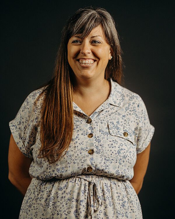 Woman with long brown hair, wearing a floral dress, smiles at the camera against a dark background.