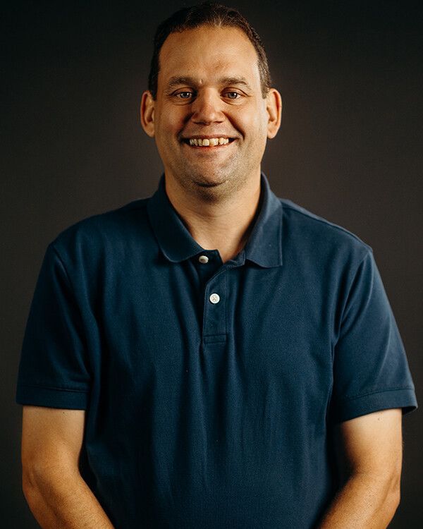 Man with short hair smiles at the camera, wearing a blue polo shirt against a gray backdrop.