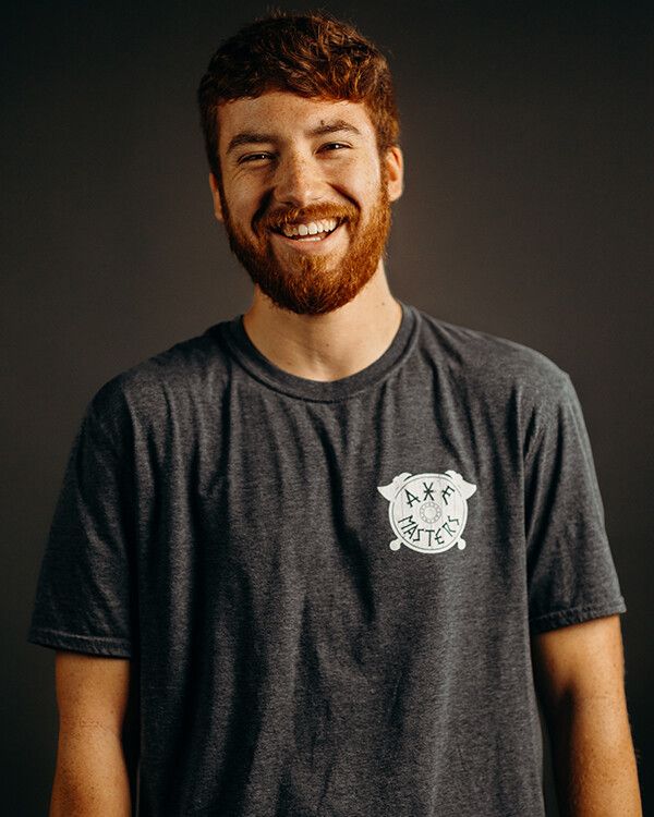 Man with red beard and hair, smiling. Wearing gray t-shirt with logo, against a dark gray backdrop.