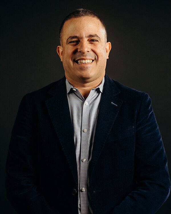 Man smiling, wearing a blue blazer over a checkered shirt, against a dark gray background.