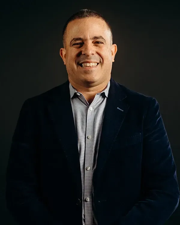 Man with a shaved head smiles, wearing a navy blazer over a patterned shirt, against a dark background.