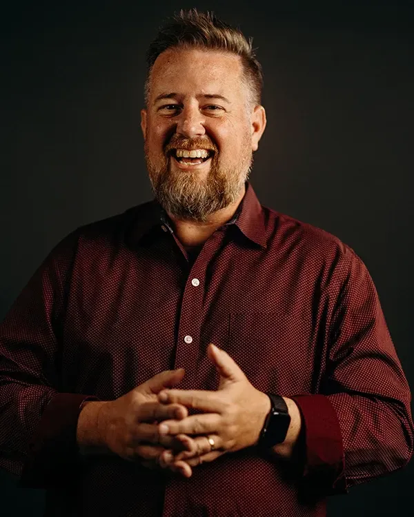Man in red shirt smiles, hands clasped. Dark background.