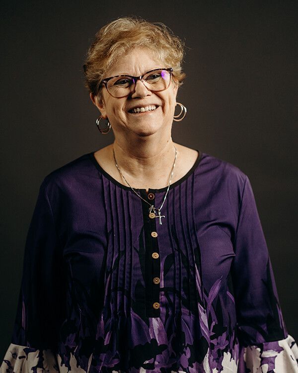 Woman in purple shirt smiles, wearing glasses and cross necklace. Gray background.