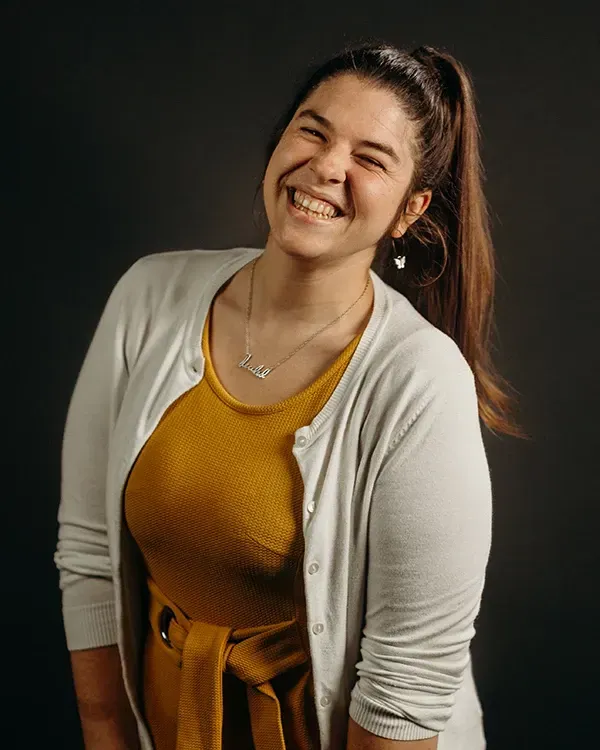 Woman smiling, wearing a mustard-colored top, grey cardigan, and high ponytail; against a dark background.