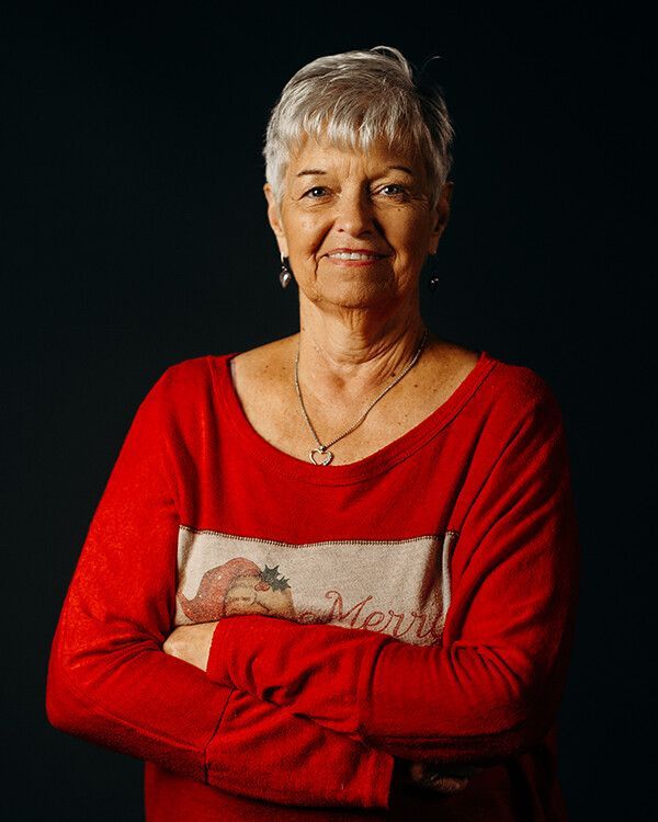 Woman with short gray hair in red shirt, arms crossed, smiling. Dark background.