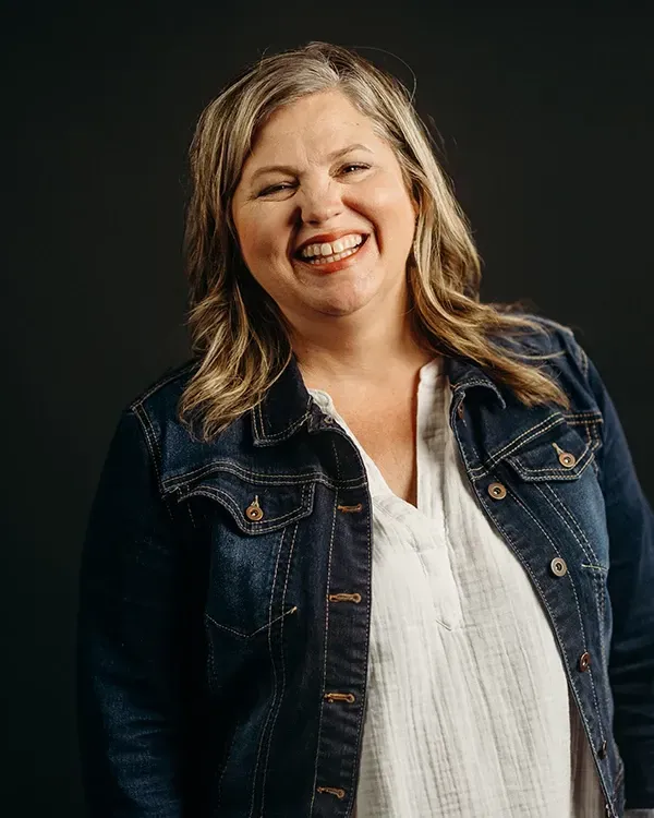 Woman with graying blonde hair smiles, wearing a denim jacket and white shirt against a dark background.