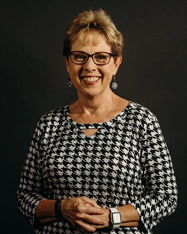 Woman in glasses and houndstooth top, smiling. Black and white pattern, dark background.