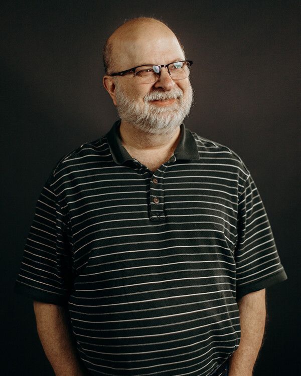 Man with glasses and a beard wearing a striped shirt, smiling against a dark background.