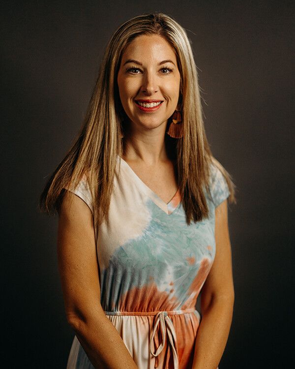 Woman with long blonde hair smiles, wearing a tie-dye dress against a dark background.