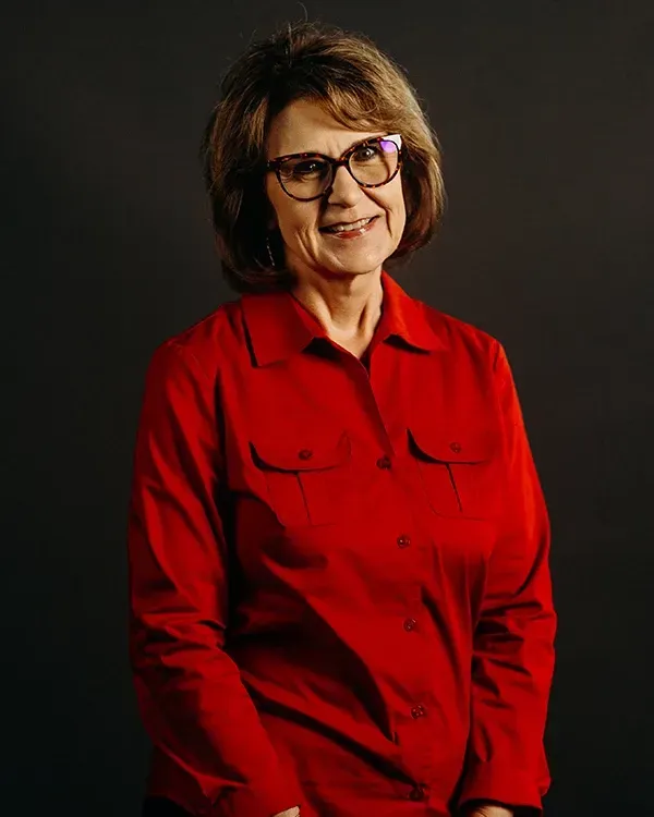 Woman in red button-up shirt and glasses smiles, posing in front of a dark backdrop.