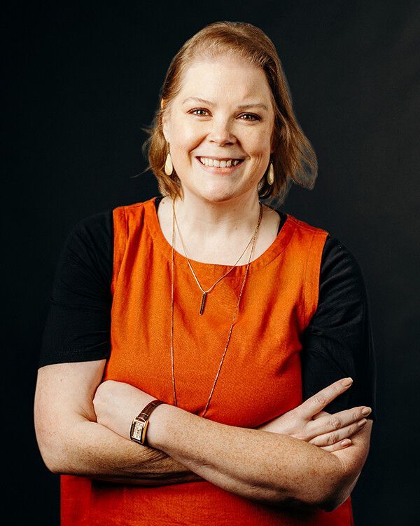Woman with crossed arms smiles, wearing an orange top with black sleeves and a gold necklace, against a dark background.