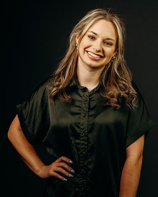 Woman in a dark green button-down shirt smiles, hand on hip, against a black backdrop.
