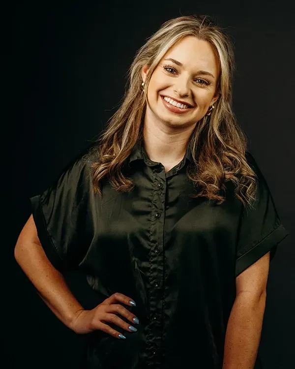 Woman smiling, hand on hip, wearing a dark green button-up shirt against a black background.