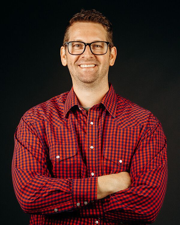 Man with glasses, arms crossed, wearing a red plaid shirt, smiling in front of a black background.